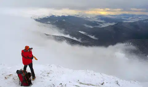 A person in a red jacket stands on a snowy mountain peak, taking a photo of the misty landscape and distant mountains at sunrise or sunset. A backpack is on the ground nearby.