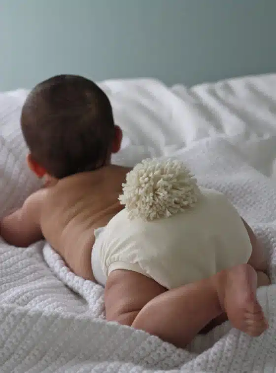 A baby lies on a white blanket, wearing a diaper with a fluffy pom-pom attached to the back.