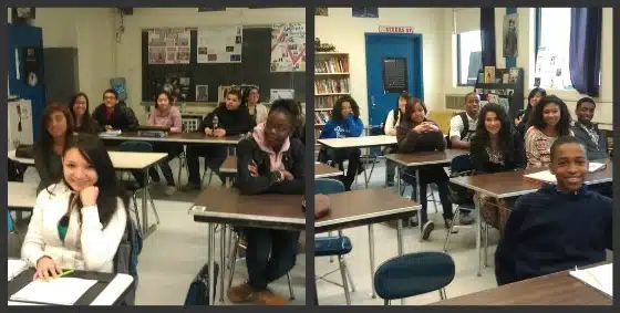 A classroom with students seated at desks, facing the camera. The room has educational posters and bookshelves in the background.
