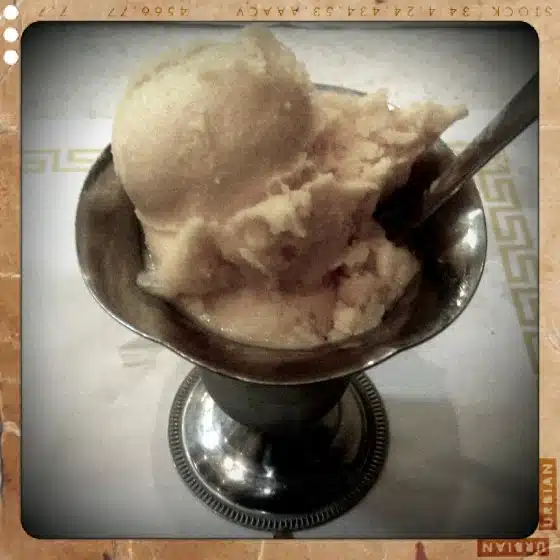 A metal dessert dish holds a scoop of light-colored ice cream with a spoon, set on a table with a white napkin and decorative edge visible.