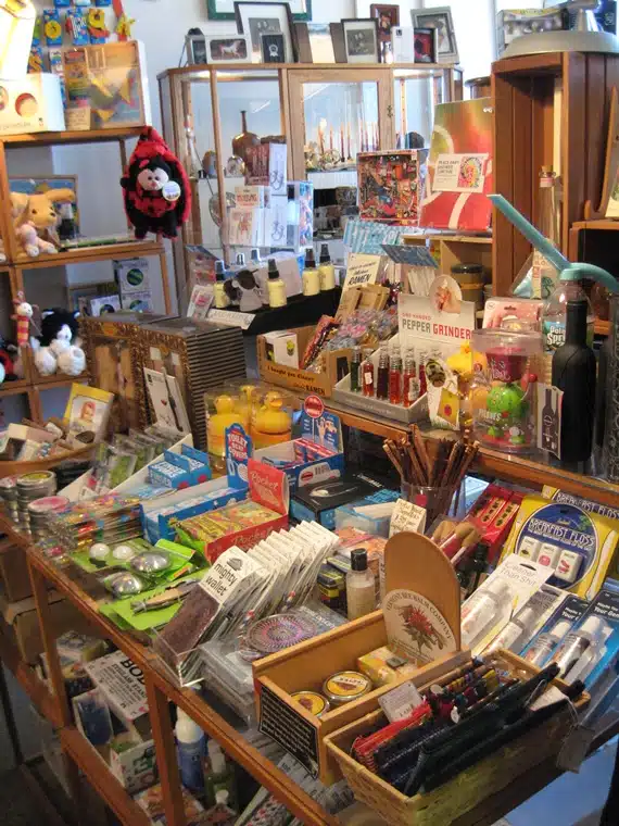 A cluttered store display featuring various small items such as toys, stationery, pepper grinders, and kitchen gadgets arranged on shelves and a central table.