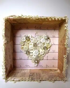 Shadow box with a heart made of white buttons on a pink background, framed by lace and vintage paper.