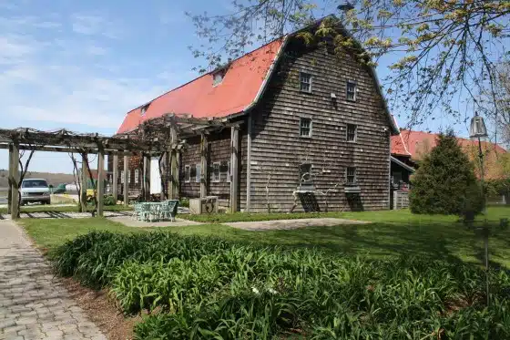 A rustic wooden barn with a red roof is surrounded by greenery, a stone path, and a sitting area with outdoor furniture. A pergola is attached to the barn, and a vehicle is parked nearby.