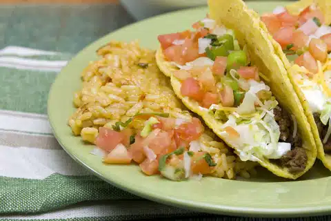 A green plate with two tacos filled with ground meat, lettuce, tomatoes, and cheese, served with a side of seasoned rice on a green and white cloth.