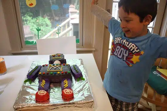 A child wearing a "Birthday Boy" shirt excitedly looks at a colorful, robot-shaped birthday cake on a table indoors.