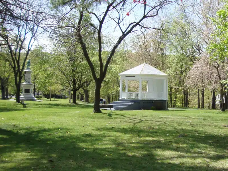 A white gazebo stands in a grassy, tree-lined park, with a small monument visible in the background under a clear sky.
