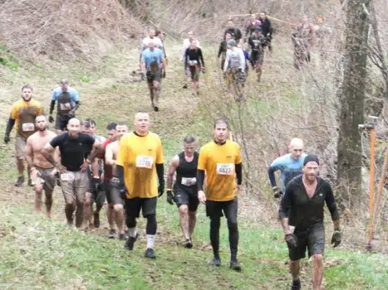 A group of runners, some wearing race bibs, navigate a muddy, uphill trail in an outdoor setting surrounded by trees and vegetation.