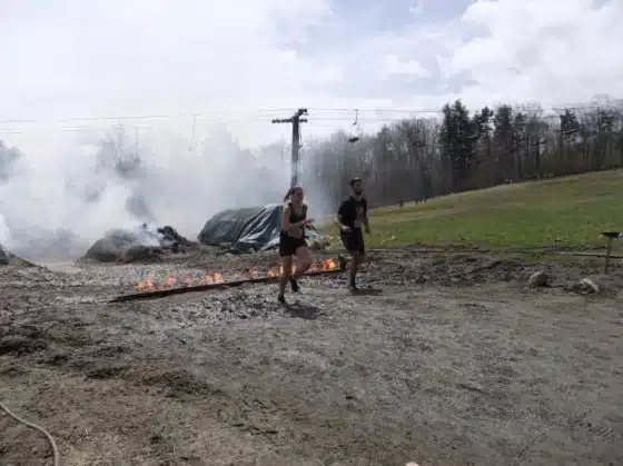 Two people are running through a muddy area with a line of fire and smoke in the background. Trees and power lines are visible in the distance.