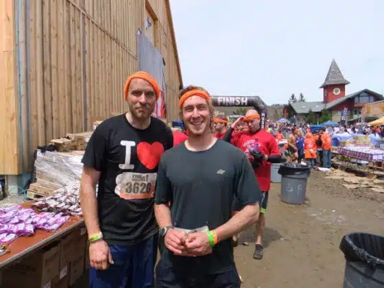 Two men wearing orange headbands stand together and smile at an outdoor event. One man is wearing a black “I ❤️” shirt and the other a dark green shirt. More participants and event structures are visible behind them.