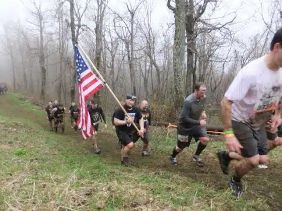 Participants in a race navigate a muddy uphill trail through a forest, with one individual carrying an American flag.