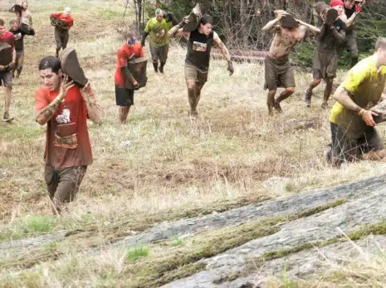 A group of individuals covered in mud are carrying large sacks on their shoulders while running up a grassy hill during an outdoor event.
