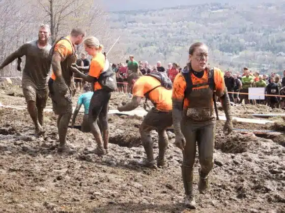 Participants in orange shirts covered in mud navigate a muddy obstacle course with onlookers in the background. Trees and a hillside are visible in the distance.