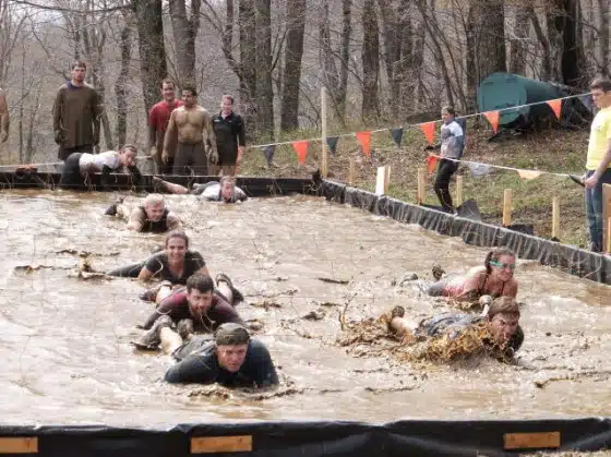 Participants crawling through a muddy obstacle course under bunting flags in a wooded area, observed by a few spectators.