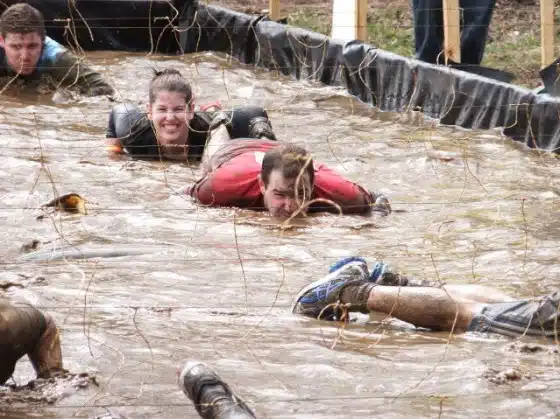 People crawl through a muddy obstacle course, navigating under barbed wire. Some individuals appear focused while one person in the foreground smiles, indicating a mix of strenuous effort and enjoyment.