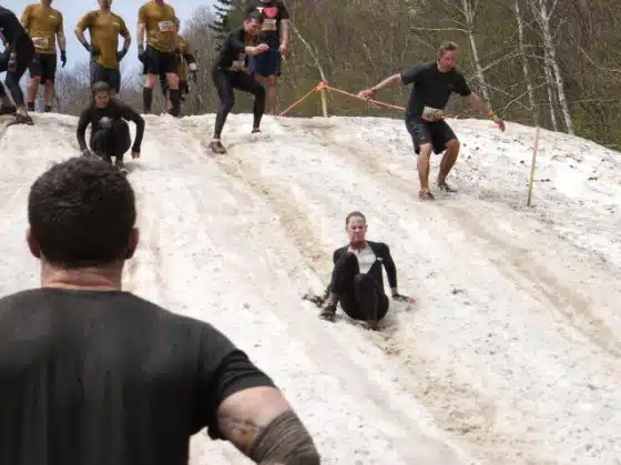 Participants slide down a muddy slope during an outdoor obstacle course race. Some are sitting while others are attempting to stand or run. Trees and ropes are visible in the background.