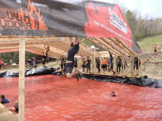 Participants navigate monkey bars over a pit of red water at the Tough Mudder event. Some are hanging, while others watch or are in the water. The atmosphere is energetic and challenging.