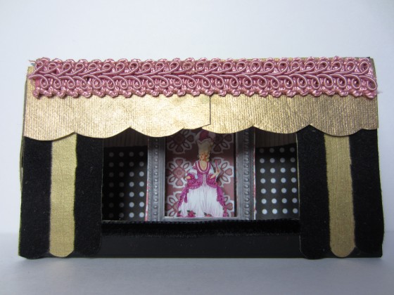 Large and small matchboxes combined to create a miniature theater. A rectangle was cut from the center of the large matchbox to create a stage. Decorative ribbon and paper was used to add detail.