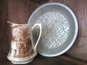 A ceramic pitcher with a brown pastoral scene stands next to a tin pie plate with a perforated pattern, both placed on a wooden surface.