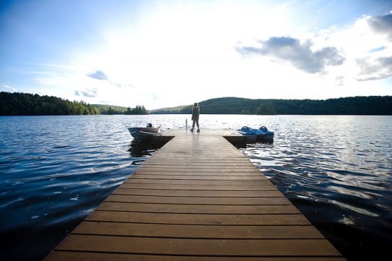 A person stands at the end of a wooden dock extending into a calm lake, with boats on either side and a backdrop of forested hills under a partly cloudy sky.