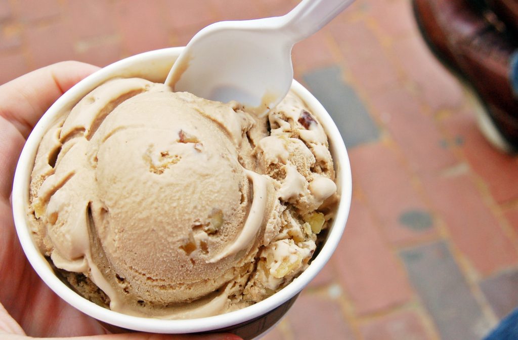 A person holds a cup of ice cream with a plastic spoon. The maple-walnut ice cream, a New Englander favorite, is served in a white paper cup. The background shows a brick pavement.