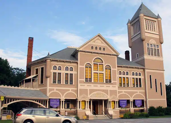 A light brown and beige historic building with a prominent tower and stained glass windows. A car is parked in front of it. Purple banners hang near the entrance.