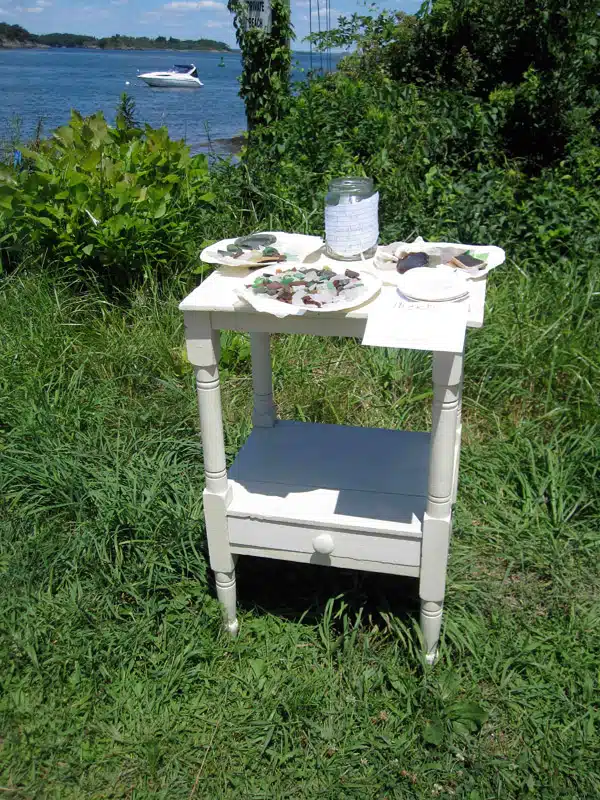 A small white table with art supplies, including paint jars and a palette, is set up outdoors on grass near a body of water with a boat visible in the background.