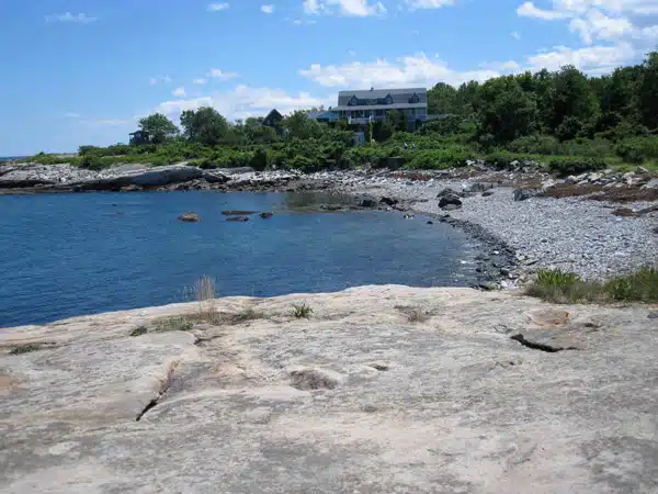 Rocky shoreline with sparse vegetation in the foreground leading to a pebble beach; a large house is situated among trees and greenery in the background under a blue sky with scattered clouds.