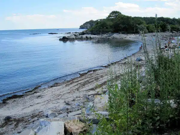 A tranquil beach scene showing calm blue water, rocky shores, and a few scattered people along the coastline with greenery in the foreground.
