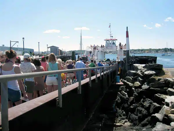 A crowd of people stands on a pier waiting to board a ferry on a sunny day. The pier is lined with a metal railing and the sea and rocky shore are visible.