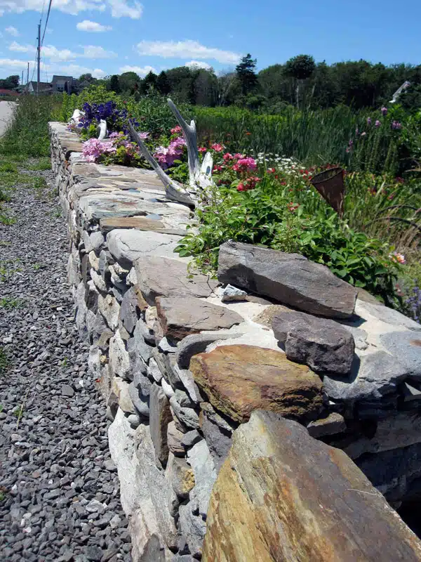 A stone wall bordering a gravel path is adorned with various plants and flowers, set against a backdrop of greenery and a clear blue sky.