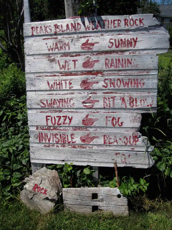 Wooden sign humorously labeled "Peaks Island Weather Rock" describes weather conditions through the state of a rock beside it, such as "Wet: Raining" and "Rock Gone: Tornado.