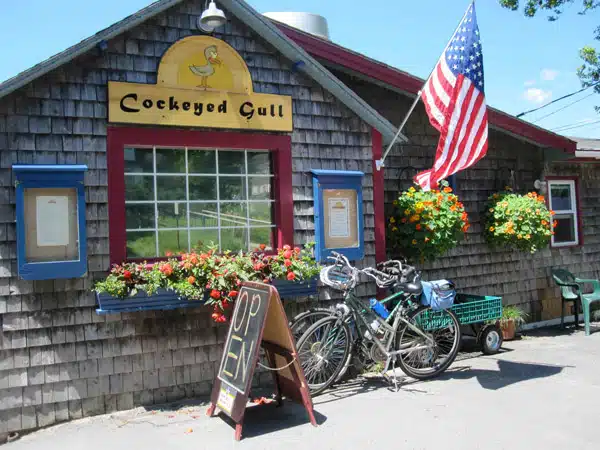 A quaint building with a sign reading "Cockeyed Gull," an American flag, two bicycles parked outside, flower baskets, and an "OPEN" board on the sidewalk.