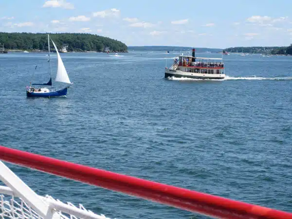 A blue sailboat and a white, red-roofed ferry boat are on a calm blue lake, surrounded by forested shores under a clear sky. A red and white railing is visible in the foreground.
