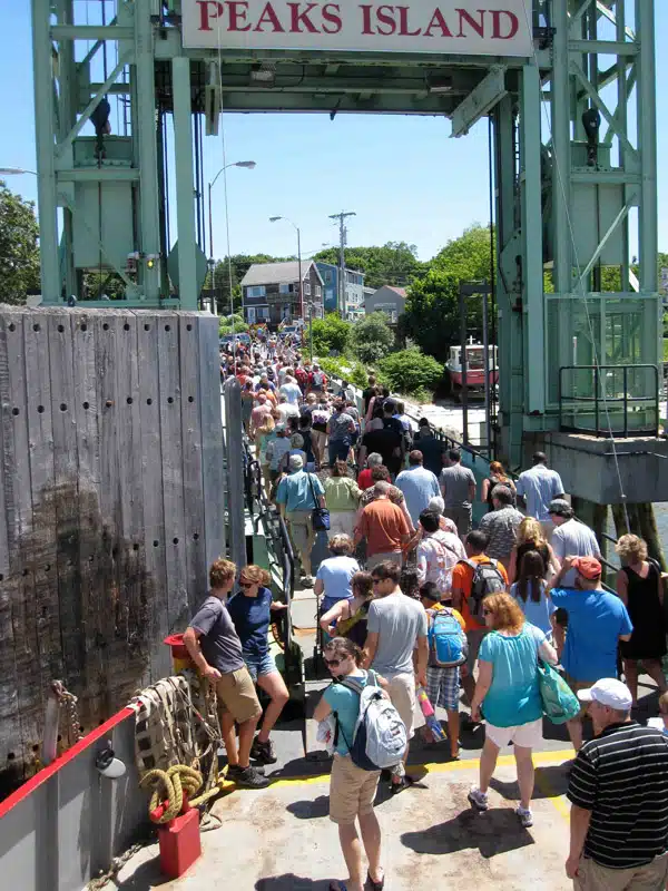 Crowd disembarking off a ferry at Peaks Island dock, with numerous people walking along a narrow gangway under a tall metal structure.