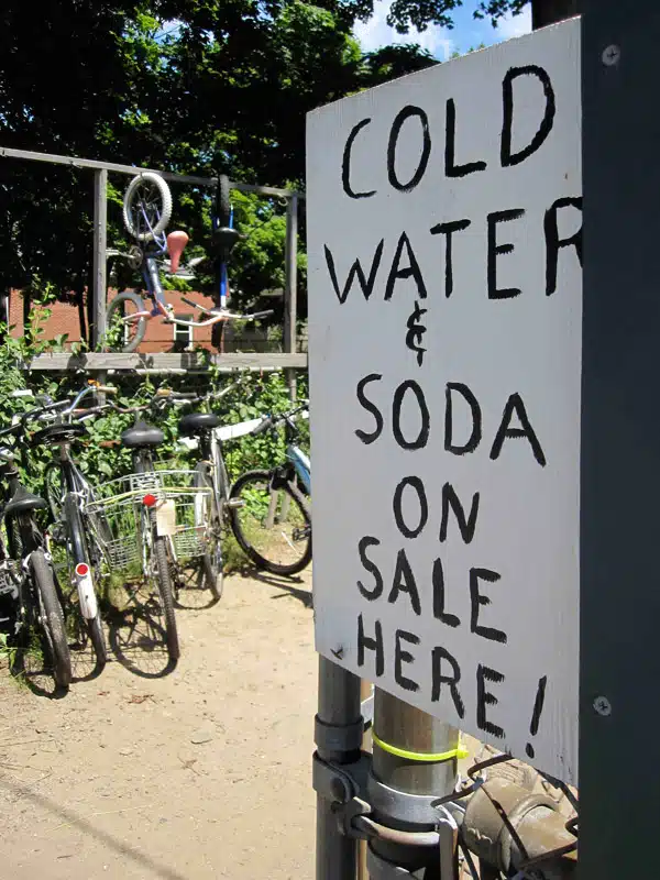 A sign reads "COLD WATER & SODA ON SALE HERE!" in front of a rack with several bicycles parked.