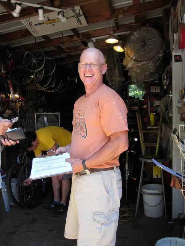 A bald man in glasses and an orange shirt stands smiling in a workshop filled with various tools and bicycles. Another man in a yellow shirt is working in the background.