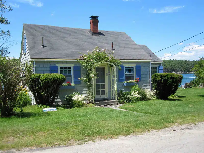Small grey house with blue shutters, a chimney, and a front yard with bushes, flowers, and a path to the door. A body of water and green trees are visible in the background.