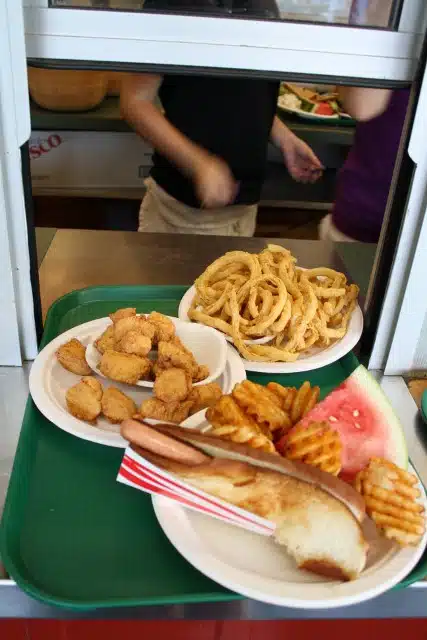 A cafeteria tray with a hot dog, waffle fries, watermelon slice, a plate of onion rings, and a plate of chicken nuggets. In the background, two people are partially visible at a service counter.