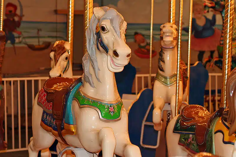 Close-up of white carousel horses with colorful saddles and bridles, against a background of a painted mural and other ride elements.