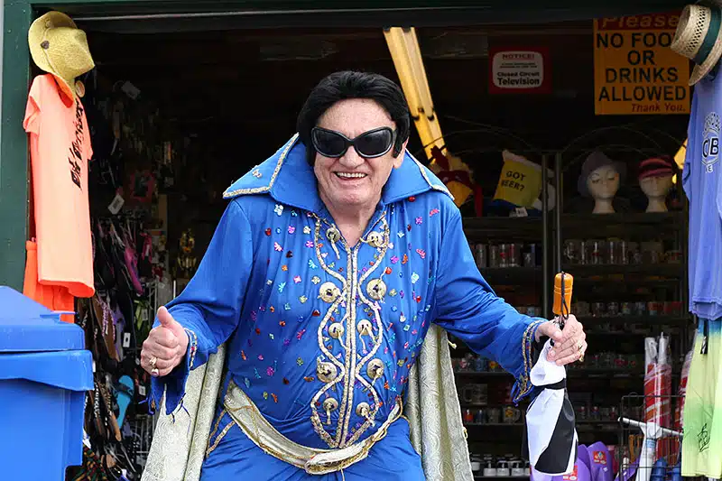 Person dressed in an Elvis Presley-inspired costume stands outside a shop with a thumbs-up gesture, holding a walking stick.