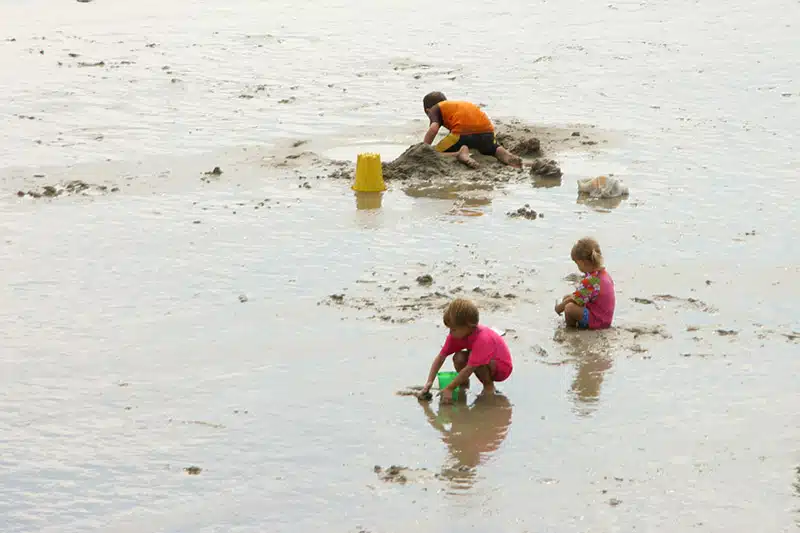 Three children play in the muddy water, building sand structures. One wears an orange shirt, another a pink shirt, and the third a red shirt. A yellow bucket is near them.