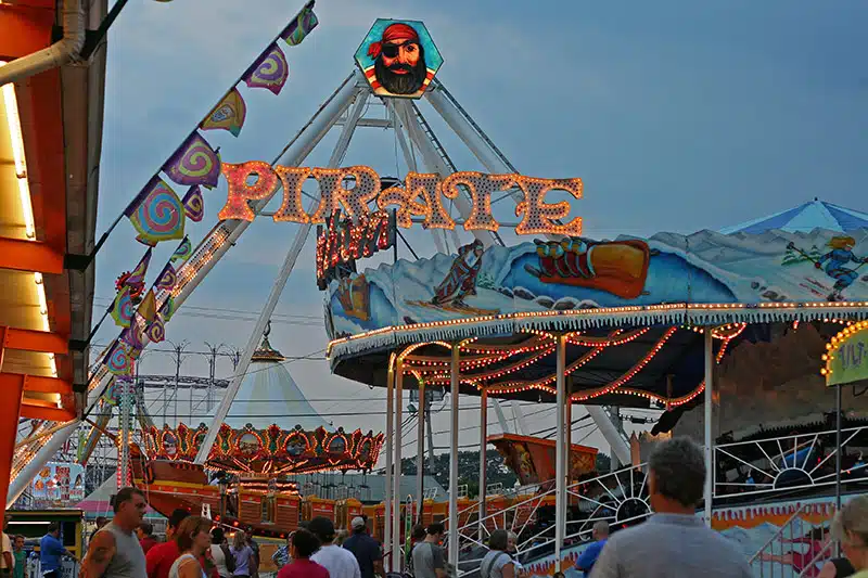 A crowd walks around a brightly lit amusement park at dusk, featuring a large "PIRATE" ride with pirate-themed decor and other colorful attractions in the background.