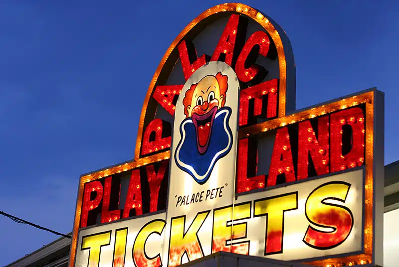 Colorful, illuminated sign reading "Playland Palace Tickets" with a clown image labeled "Palace Pete" against a twilight sky.