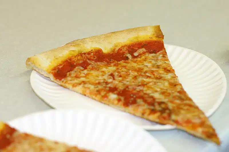 A slice of cheese pizza on a white paper plate is placed on a light-colored table surface.