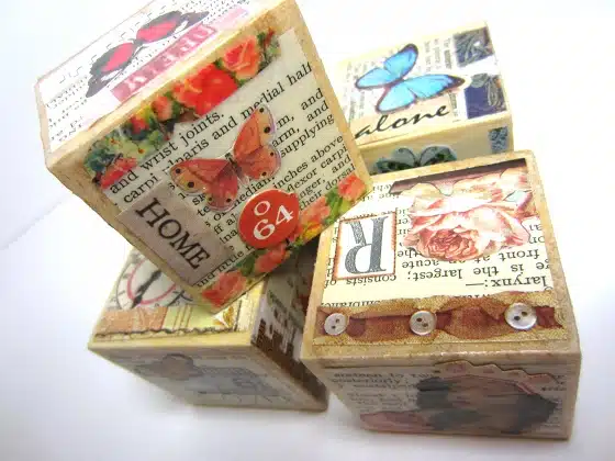 Four decorative wooden blocks with various colorful collage designs including butterflies, flowers, text, and images are stacked together against a white background.