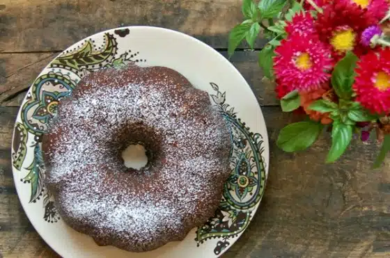 A bundt cake dusted with powdered sugar sits on a decorative plate next to a small vase of pink and red flowers on a wooden table.