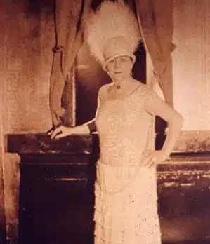 A sepia-toned image of a woman wearing a decorative hat with white feathers, a light-colored patterned dress, and long necklace, standing in front of a dark background with curtains.