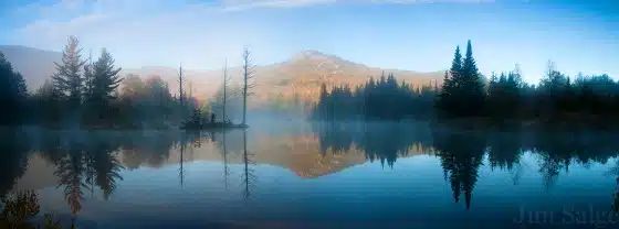 A serene lake with a misty surface reflects the surrounding forest and distant mountains under a clear sky.