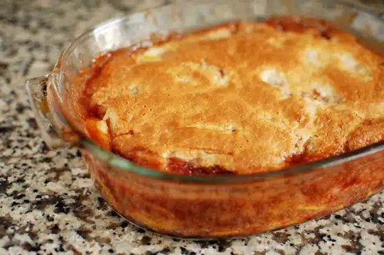 A baked dish with a golden brown crust, likely a peach cobbler, in a clear glass baking dish placed on a speckled countertop.