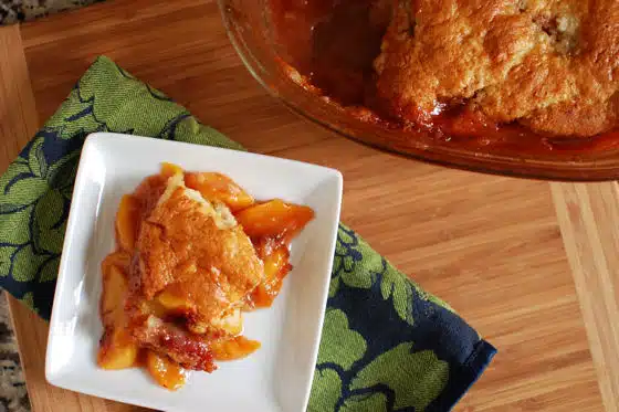 A serving of peach cobbler on a white square plate with a glass baking dish containing the remaining cobbler, placed on a wooden surface with a green and blue cloth.